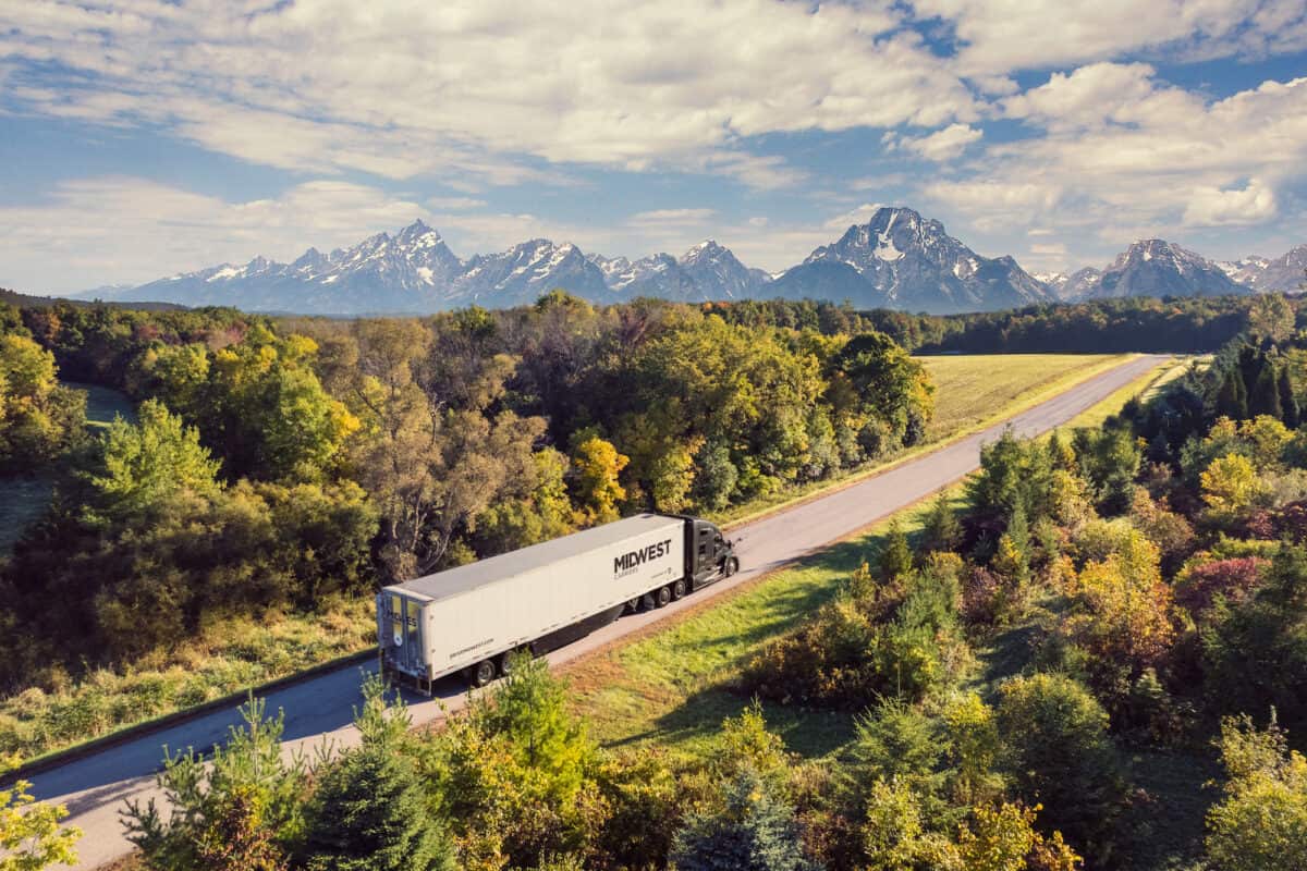 Midwest Carriers truck drives along road with trees surrounding the area and mountains in the background