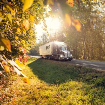 A Midwest Carriers truck on the highway with the sun shining above the truck and fall leaves on the side of the road.