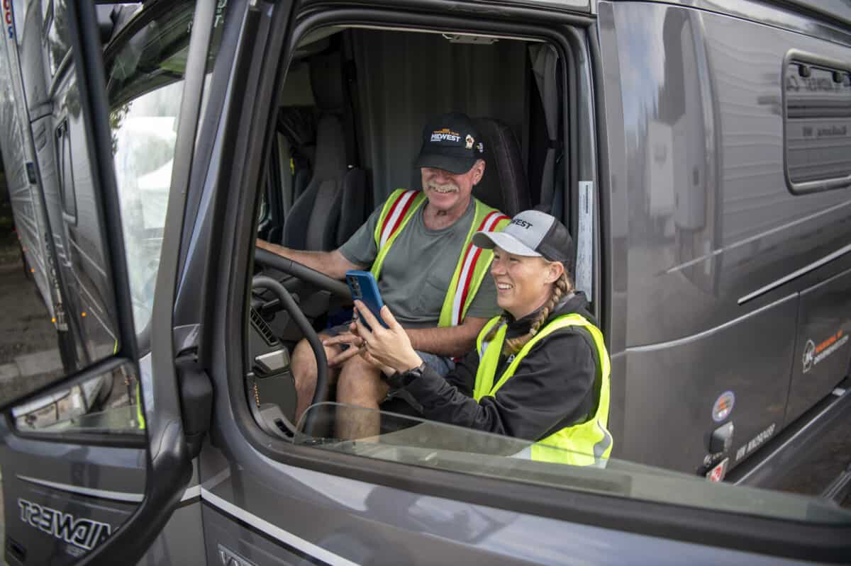 One Midwest Carriers driver sits in behind the wheel of a   semi, while another stands beside them, pointing to features within the cab of the truck.