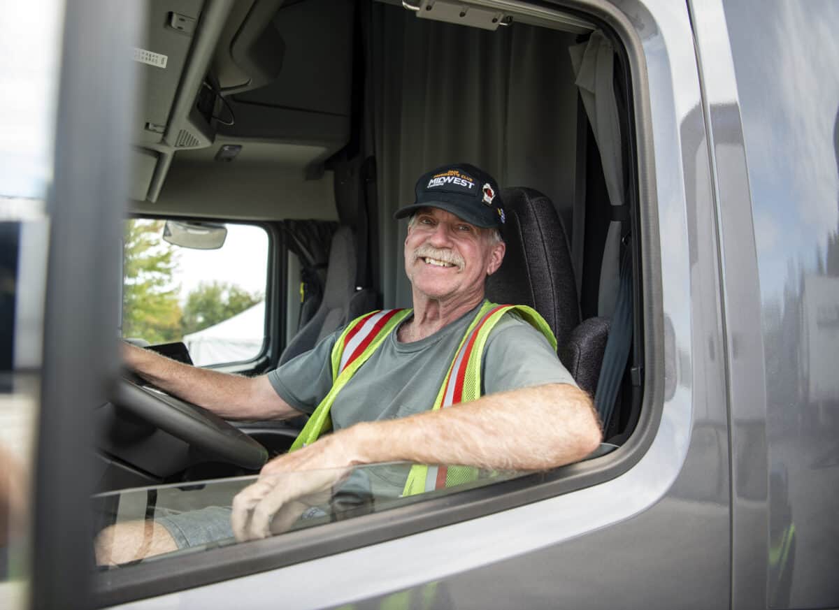 A Midwest truck driver wearing a baseball cap smiles at the camera  from behind the wheel of his truck.