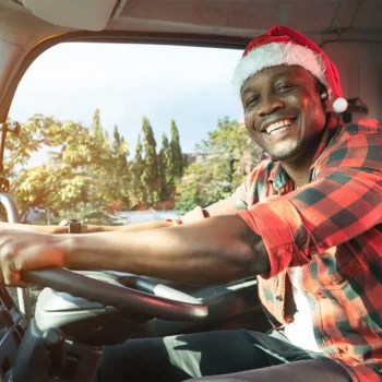 A male truck driver in plaid clothing driving a truck wearing a santa hat.