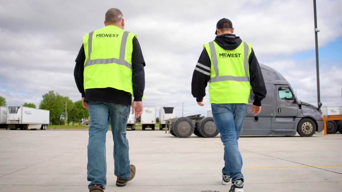 Two Midwest Carriers employees wearing safety vests walking across the yard toward a semi-truck