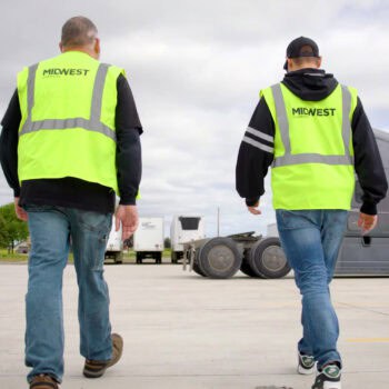 Two Midwest Carriers employees wearing safety vests walking across the yard toward a semi-truck